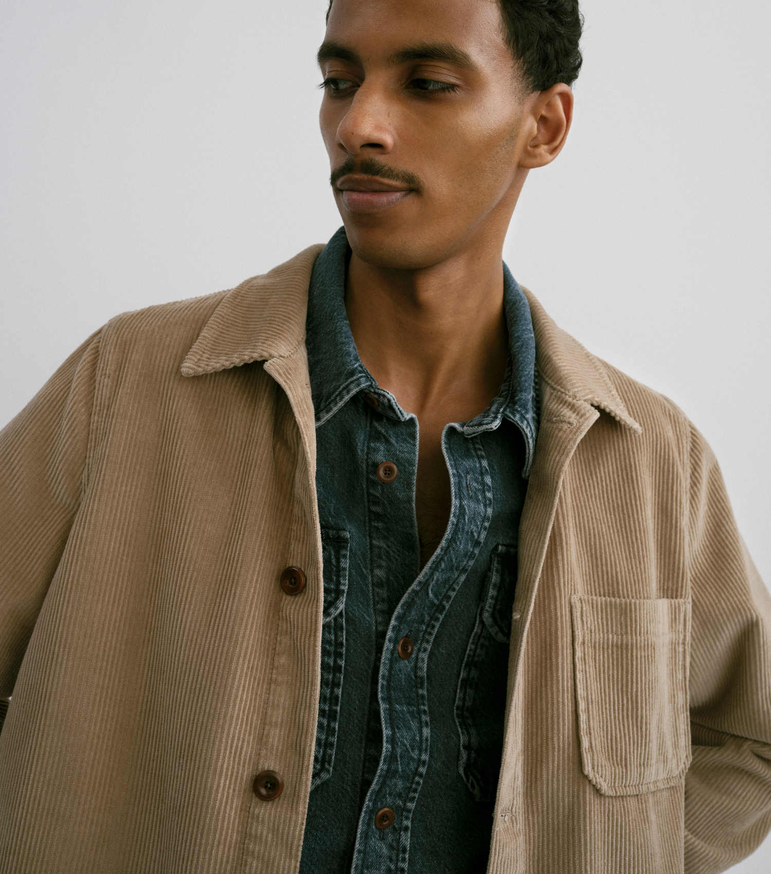 Man wearing a beige corduroy jacket over a denim shirt against a plain background