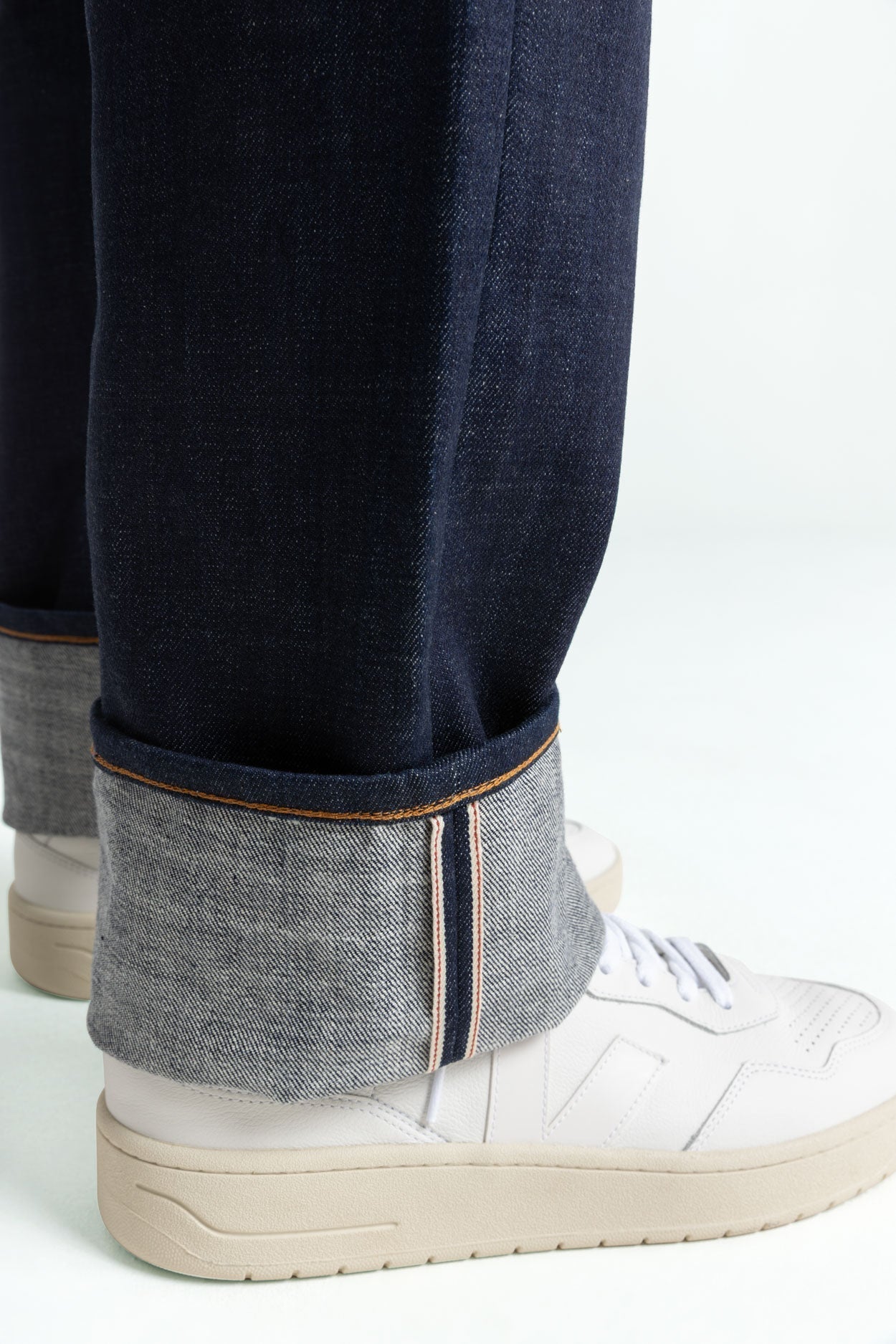 Close-up of a pair of dark raw denim jeans with a wide cuff, revealing the selvedge edge with contrast stitching. The wearer pairs the jeans with clean white sneakers, showcasing a classic and minimalistic style.