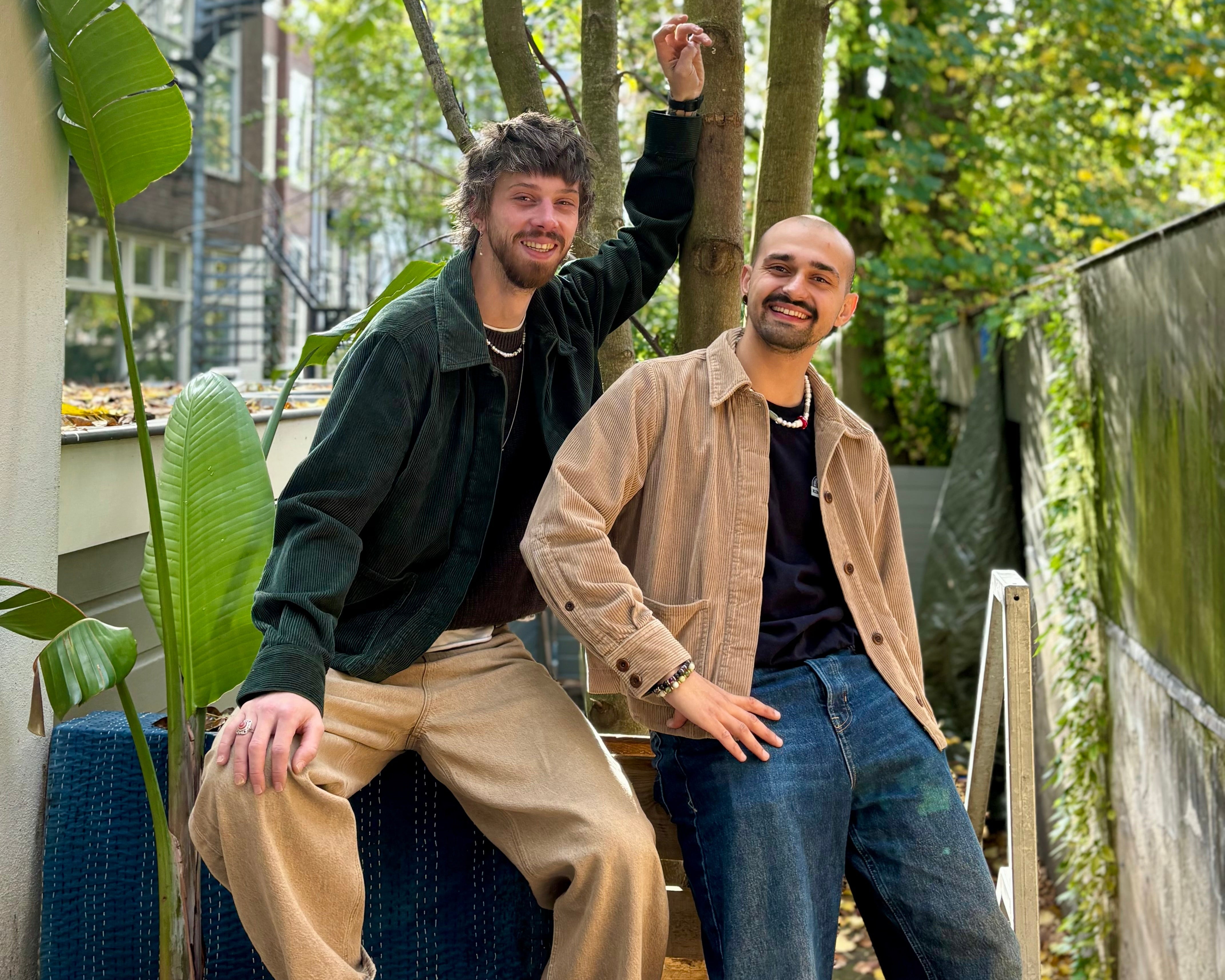 Two men posing outdoors with trees and buildings in the background